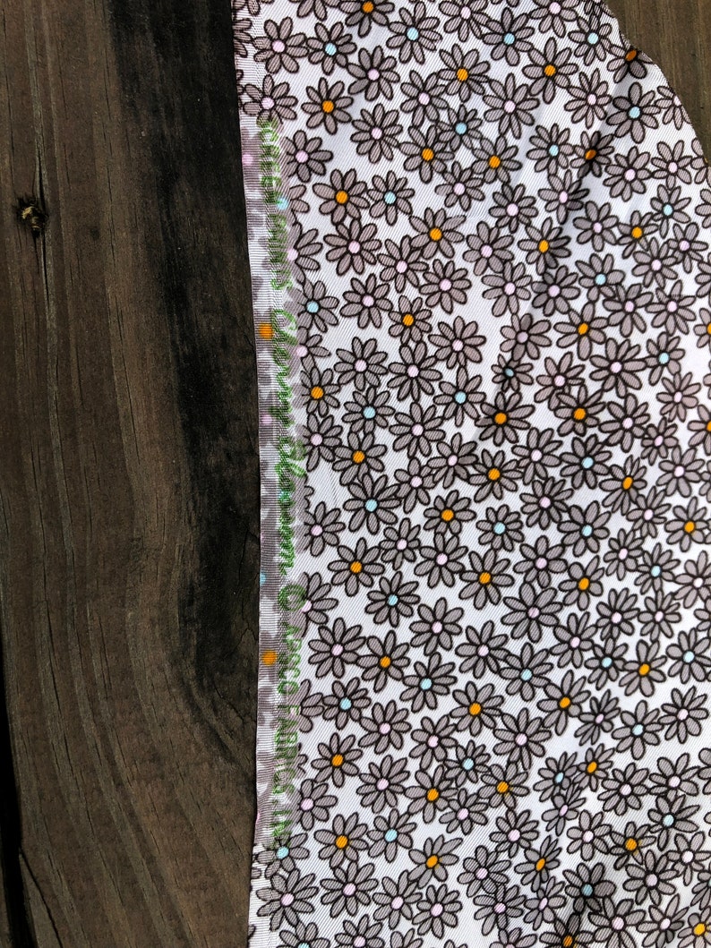 Puede incluir: Primer plano de una tela con fondo blanco cubierta de peque&ntilde;as flores de margarita grises con centros naranjas y celeste. El borde de la tela tiene un borde verde y blanco con texto.