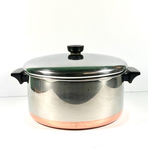 May include: A stainless steel cooking pot with a copper-colored base and a matching lid. The pot features two black side handles and a small black knob on the center of the lid. The exterior surface is reflective and smooth with a cylindrical shape.