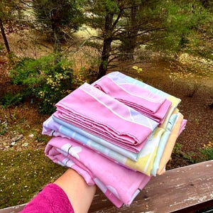 May include: A stack of folded, pastel-colored towels. The towels are primarily pink, with accents of blue, yellow, and purple. The towels are held in front of a natural outdoor background with trees and foliage.