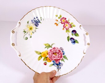 Op de afbeelding: Witte ovale serveerschaal met gouden rand en bloemmotieven. De schaal heeft twee handvatten en heeft een verscheidenheid aan bloemen, waaronder roze pioenen, gele citroenen en blauwe vergeet-mij-nietjes.