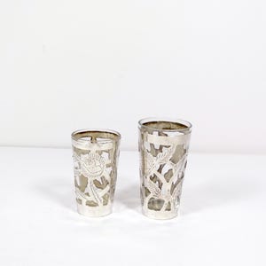 May include: Two silver-plated drinking glasses with cut-out floral designs. The glasses are clear and the designs are intricate.