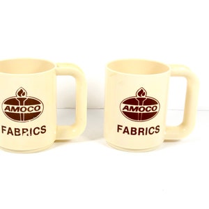 May include: Two white ceramic mugs with brown Amoco Fabrics logos. The logos feature a flame and the Amoco name in a brown oval.