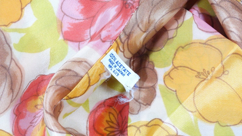 Pode incluir: Close-up de um tecido branco com estampa floral em tons de laranja, amarelo e verde. O tecido tem uma etiqueta que diz "100% Acetato Feito no Jap&atilde;o HPL 6119".