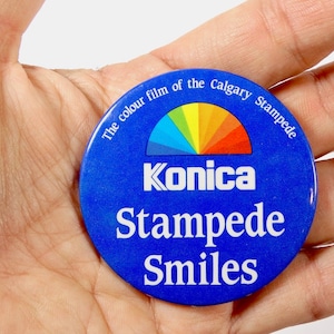 May include: Blue and white button with the text "The colour film of the Calgary Stampede" and "Konica Stampede Smiles". A rainbow arc is above the text "Konica".