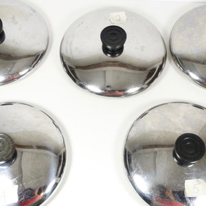 May include: Five silver pot lids with black knobs. The lids are arranged in a semi-circle on a white surface.