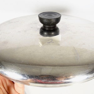 May include: A stainless steel pot lid with a black knob. The lid is round and has a shiny, reflective surface. A small piece of paper with the number 2 is attached to the lid.