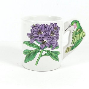 May include: White ceramic mug with a purple rhododendron flower design and a green hummingbird handle.