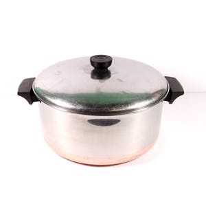 May include: A stainless steel cooking pot with a copper-colored base, featuring two black side handles and a matching stainless steel lid with a black knob. The pot has a cylindrical body and a polished metallic finish on the exterior surface.