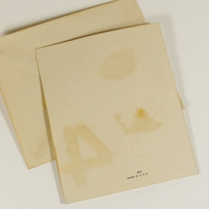 May include: A blank greeting card with a white background and a light brown envelope. The card has a faint brown stain on the back. The envelope is slightly folded. The card has the text "Made in U.S.A." printed on the back.