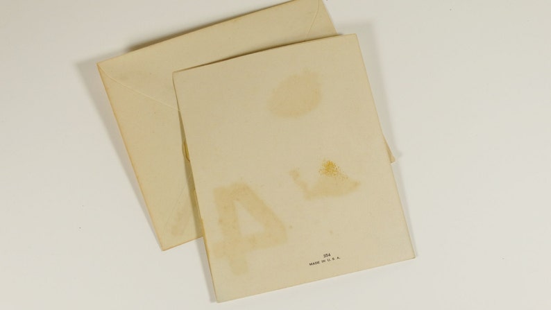 May include: A blank greeting card with a white background and a light brown envelope. The card has a faint brown stain on the back. The envelope is slightly folded. The card has the text "Made in U.S.A." printed on the back.
