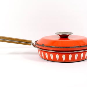 May include: A vintage orange enamel pan with a lid and wooden handle. The pan features a white leaf pattern around the base and a silver rim. A classic kitchenware item.