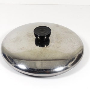 May include: A silver pot lid with a black knob. The lid is round and has a smooth surface.