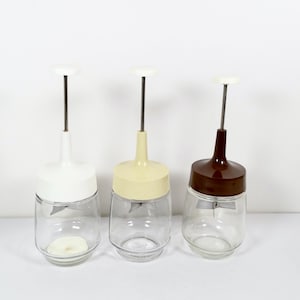 May include: Three glass food mills with different colored plastic tops: white, cream, and brown. The mills have a metal blade inside and a plunger on top.