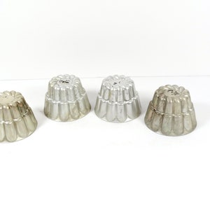 May include: Four silver metal jelly molds with a fluted design. The molds are arranged in a row on a white surface.