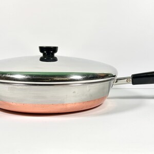 May include: A round stainless steel skillet with a copper-colored base and a matching lid. The pan features a black handle with a small metal loop at the end and a black knob on the center of the lid. The exterior has a polished metallic finish.