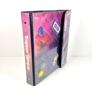 May include: A Mead Trapper Keeper binder featuring a dark purple and pink galaxy-themed cover with graphic prints of a cassette tape, palm trees, and the text OMG!. The spine is dark blue with the white text Trapper Keeper printed vertically.