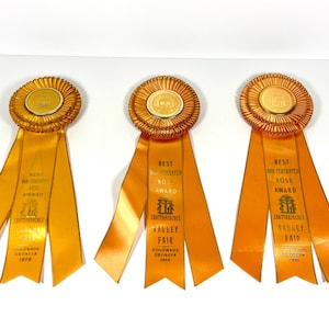 May include: Three orange satin rosette ribbons with gold-tone circular centers. Each ribbon features three hanging tails printed with the text: Best Non-Federated Rose Award, Chattahoochee Valley Fair, Columbus Georgia, and the years 1976, 1988, and 1990.