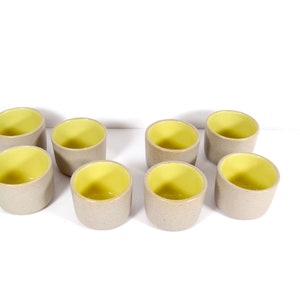 May include: Eight small ceramic cups with a light gray exterior and a bright yellow interior. The cups are arranged in two rows of four.