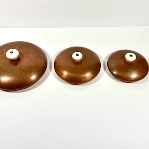 May include: Three circular copper lids of varying sizes, each featuring a smooth, metallic finish and a small, centered white ceramic knob. The lids are arranged in a horizontal row from largest to smallest, showcasing a consistent design.