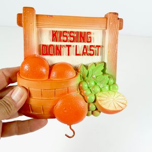 May include: A vintage orange-themed wall decor with a sign that reads "Kissing Don't Last". The decor features a basket of oranges, grapes, and a lemon slice.