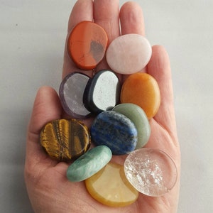 May include: A hand holding a collection of polished gemstones in various colors and textures. The stones include red jasper, rose quartz, tiger's eye, lapis lazuli, and clear quartz, among others. The stones are smooth and oval-shaped.