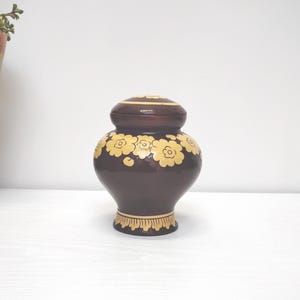 Könnte beinhalten: Eine dunkelbraune dekorative Urne mit Deckel, verziert mit einem sich wiederholenden Muster aus cremefarbenen floralen Designs. Die Urne hat einen abgerundeten Körper und einen dekorativen Sockel. Der Deckel hat auch ein cremefarbenes Blumendesign.