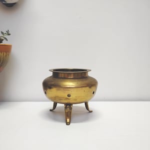 May include: A round brass vessel with a wide opening and a smooth, metallic finish. The container features three short, curved legs and a series of small, evenly spaced circular holes positioned around the midsection of the body.