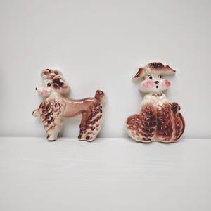 May include: Two ceramic poodle figurines. One is standing, the other is sitting. Both are primarily brown with pink accents on the ears and face. The standing poodle is approximately 4 inches long. The sitting poodle has rosy cheeks and a small collar.