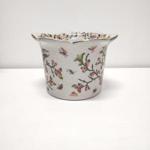 May include: A white ceramic planter with a scalloped rim and a crackle glaze finish. The exterior is decorated with colorful illustrations of various insects, including butterflies and a snail, interspersed with flowering branches and buds.