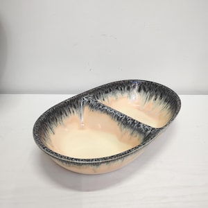 Può includere: Un piatto da portata ovale in ceramica con un divisorio centrale. L'interno presenta una smaltatura liscia color pesca pallido, mentre il bordo esterno è rifinito con un motivo a goccia screziato nei toni del nero e del grigio scuro.