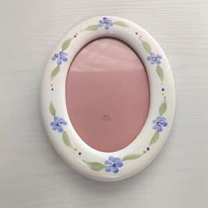 May include: An oval white ceramic picture frame featuring a hand-painted floral pattern of blue flowers and green leaves along the border. The center of the frame contains a pink insert with the printed text "SIZE 5" x 7"" in the middle.