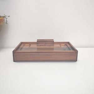 May include: A rectangular wooden desk organizer featuring a dark brown finish with a smooth grain pattern. The piece includes a central raised tiered stand, two circular recessed compartments on either side, and a flat base with raised edges.