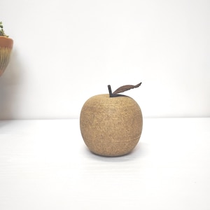 Könnte beinhalten: Eine dekorative Apfelskulptur mit einer strukturierten, hellbraunen Oberfläche. Der Apfel hat einen kleinen, dunkelbraunen Stiel und ein Blatt. Die Skulptur steht vor einem weißen Hintergrund, was auf eine minimalistische Ästhetik hindeutet.