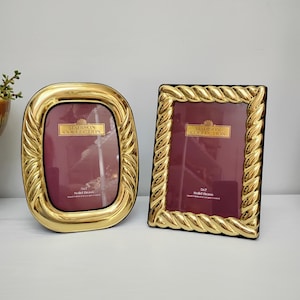 May include: Two Madison Collection picture frames made of solid brass with a polished gold finish. One frame is oval with a smooth, rounded border, and the other is rectangular with a twisted rope-style edge. Both frames hold 5x7 inch inserts.