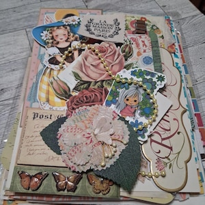 May include: A layered paper craft collage featuring vintage-style illustrations of a girl in a blue hat, a rose, and a cartoon character. The composition includes a fabric flower, a string of cream-colored beads, and three butterfly cutouts.