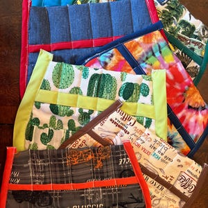 May include: Assortment of colorful quilted aprons with various patterns, including cacti, denim, and coffee-themed designs. Each apron features a different trim color and pocket design. The aprons are made of fabric.