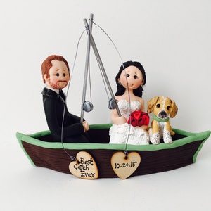 Puede incluir: Un adorno para pastel de boda con una novia y un novio en un bote marrón y verde con cañas de pescar. La novia lleva un ramo de flores rojas y el novio lleva una caña de pescar. El bote tiene un borde verde y una base de madera marrón. El adorno para pastel también incluye un perro pequeño sentado en el bote. El bote tiene dos letreros en forma de corazón colgando, uno dice "Best Catch Ever" y el otro dice "10-24-15".