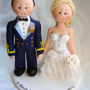 Può includere: Un topper per torta nuziale con una figurina di sposa e sposo. Lo sposo indossa un'uniforme militare blu con bottoni dorati e un papillon nero. La sposa indossa un abito bianco con un corpetto di perline e una gonna a balze. Il topper per torta è su una base di torta bianca con l'iscrizione "Shawn & Melanie July 13, 2013".