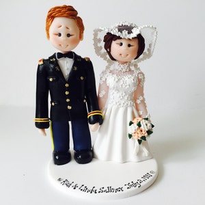 Può includere: Un topper per torta nuziale bianco con una figurina di sposa e sposo. Lo sposo indossa un'uniforme militare blu con bottoni dorati e la sposa indossa un abito bianco in pizzo con velo. Il topper per torta è su una base bianca con l'iscrizione "Rich & Linda Sellner July 23, 1981."