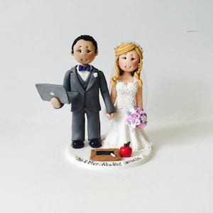 Può includere: Un topper per torta nuziale con una figurina di sposo e sposa. Lo sposo tiene un laptop e la sposa tiene un bouquet di fiori rosa e viola. La base del topper è decorata con una lavagna, una mela e il testo "Mr. & Mrs.  Adamit 01/24/16".