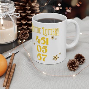 May include: White ceramic mug with the text "WIN THE LOTTERY" in gold lettering. The mug features the numbers 461, 05, and 67, along with star accents. The mug is filled with a dark liquid.