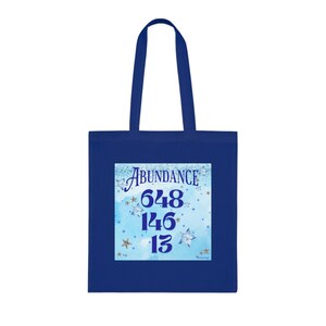 May include: A blue tote bag with a white square design featuring the word "Abundance" and the numbers 648, 146, and 13. The design is surrounded by silver stars on a light blue background.