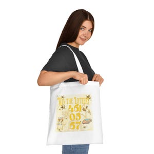 May include: White tote bag featuring a gold design with the text "Win the Lottery". The design includes the numbers 461, 05, and 67, alongside illustrations of stars, champagne flutes, and a sports car.