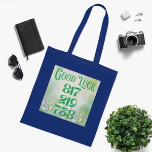May include: A navy blue tote bag with a rectangular design that reads "Good Luck" and features the numbers 817, 219, and 738 in green. The design is set against a pastel floral background. The bag has two navy blue handles.