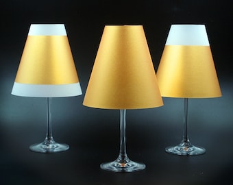 May include: Three gold and white lampshades with a cone shape, each attached to a clear glass wine glass base.