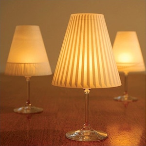 May include: Three white lampshades with vertical stripes are placed on wine glasses, creating unique table lamps. The lamps are lit and casting a warm glow.