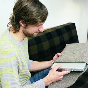 Puede incluir: Una persona sentada en un sofá con un suéter a rayas y jeans, sosteniendo una funda de computadora portátil gris con una computadora portátil dentro.