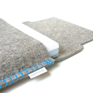 Puede incluir: Una funda para portátil de fieltro gris con costuras azules y el texto "Apfelnasche" en una etiqueta blanca. La funda está doblada para mostrar el interior.