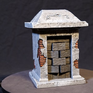 Op de afbeelding: Een miniatuur stenen pilaar met een verweerde uitstraling. De pilaar heeft een grijze, getextureerde buitenkant met zichtbare bakstenen secties. De bovenkant heeft een decoratief bloemmotief. De pilaar is een model of rekwisiet.