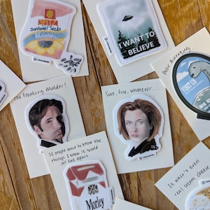 May include: A selection of stickers displayed on a wooden surface. The stickers showcase diverse designs, such as a UFO, the phrase "I WANT TO BELIEVE", a pack of Morley cigarettes, and character portraits. Other stickers include a bag of sunflower seeds and the text "trust no one".